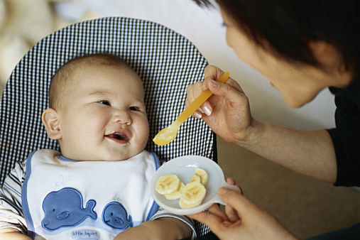 Mother feeding baby boy (9-12 months) bananas, baby smiling